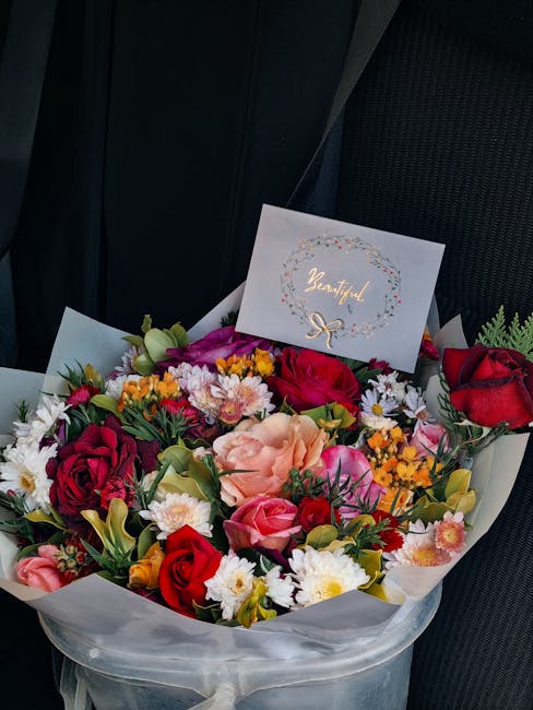 A vibrant, fresh bouquet arrangement featuring red roses, pink roses, peach carnations, yellow and white daisies, and purple statice, all artistically assembled with green foliage. The bouquet is wrap