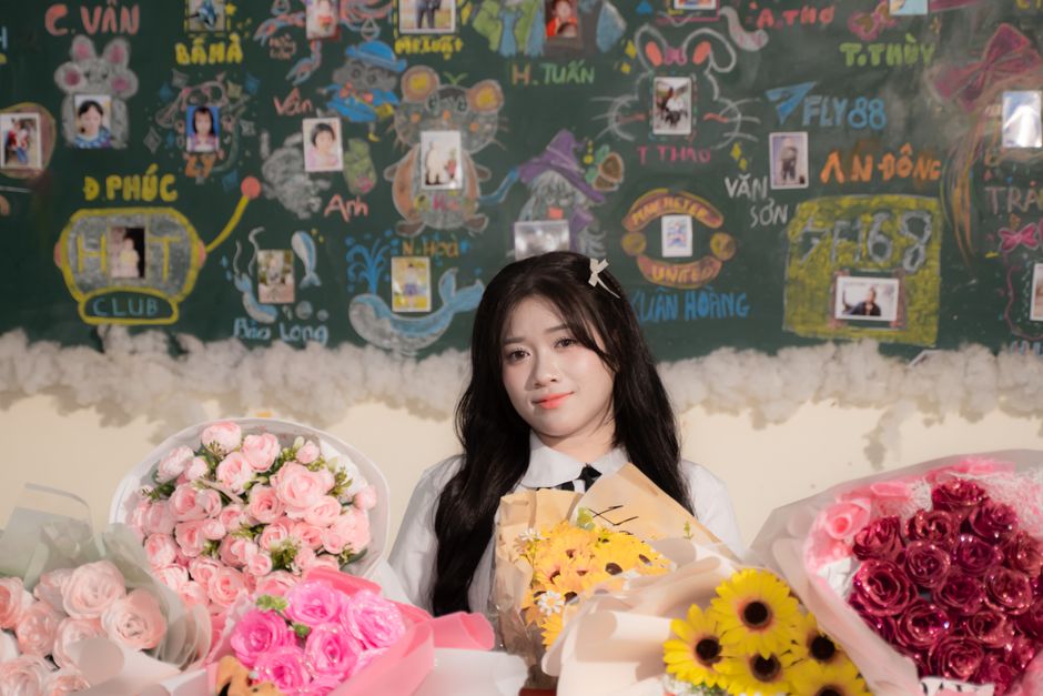 A young woman with long dark hair wearing a white blouse and a hair accessory, smiling softly, is surrounded by an array of fresh floral bouquets displayed on a table in front of her. The bouquets inc