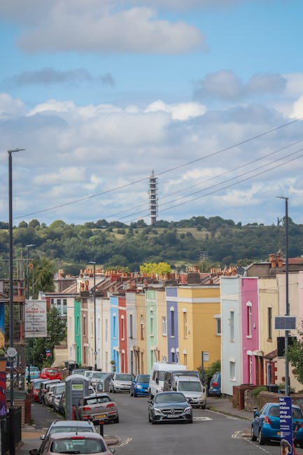 A row of colorful, pastel-painted terraced houses on a residential street, with potted plants and small trees in front gardens. Parked and moving cars line both sides of the street, while overhead pow