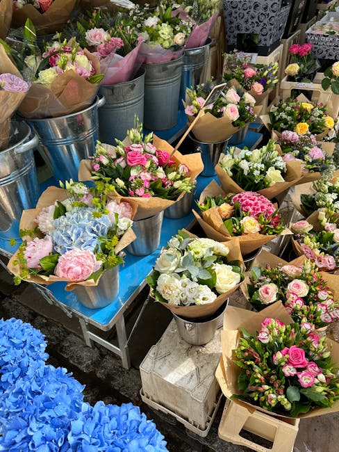 A variety of fresh floral bouquets arranged on tables at a flower market, featuring soft pink, white, and cream roses, hydrangeas, and other blossoms wrapped in brown craft paper, displayed in metalli